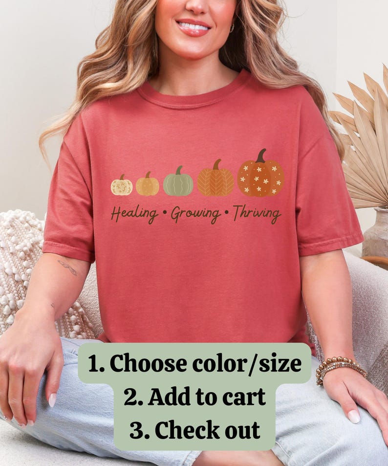 Puede incluir: Una camiseta de color rojo anaranjado con una fila de cinco calabazas de varios colores y dise&ntilde;os. El texto "Healing &bull; Growing &bull; Thriving" est&aacute; impreso debajo. Debajo del texto est&aacute;n las palabras "1. Choose color/size 2. Add to cart 3. Check out".
