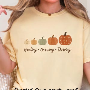 Puede incluir: Camiseta amarillo claro con una fila de cinco calabazas de varios tama&ntilde;os y colores. El texto "Healing &bull; Growing &bull; Thriving" est&aacute; debajo de las calabazas. La frase "Created by a psych. prof." est&aacute; en la parte inferior.