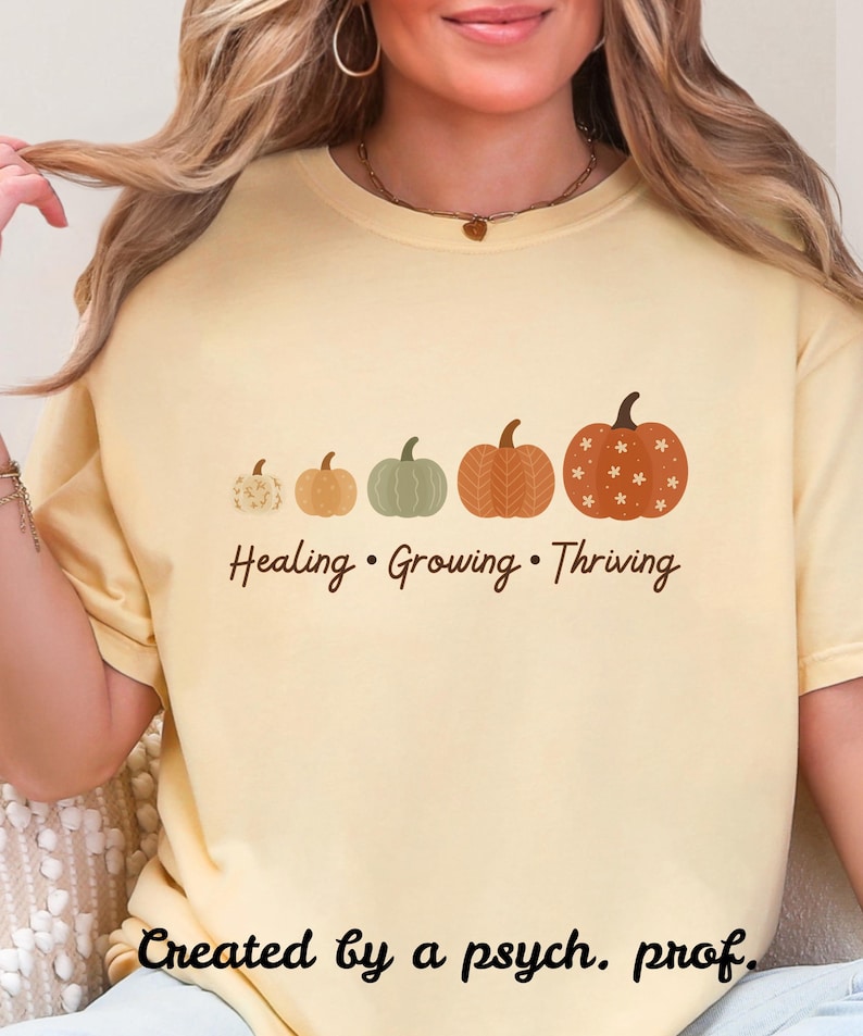 Puede incluir: Camiseta amarillo claro con una fila de cinco calabazas de varios tama&ntilde;os y colores. El texto "Healing &bull; Growing &bull; Thriving" est&aacute; debajo de las calabazas. La frase "Created by a psych. prof." est&aacute; en la parte inferior.