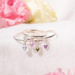 May include: Four silver rings with heart-shaped gemstone charms in blue, pink, purple, and green. The rings are displayed on a white surface with pink and yellow flowers in the background. The gemstones are small and delicate.