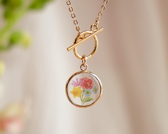 May include: A gold chain necklace with a toggle clasp and a round pendant featuring a pressed flower design. The pendant is clear and shows a variety of colorful flowers.