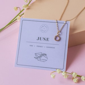 May include: A purple gemstone necklace with a gold chain and a card that says "June" with the words "Kind, Friendly, Humorous" underneath. The card also has a small illustration of a crystal.
