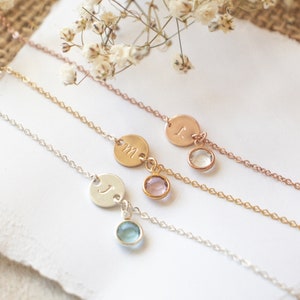 May include: Three delicate gold necklaces with initial charms and small gemstone accents. The necklaces are layered on a white surface with a sprig of white flowers.