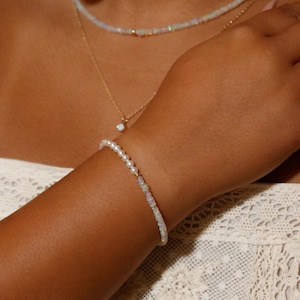 May include: A delicate bracelet featuring a strand of small white pearls and iridescent opal beads. The bracelet is worn on the wrist, complemented by a matching opal beaded necklace and a thin gold chain necklace with a single opal pendant.
