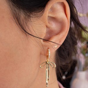 May include: Gold hoop earrings with a dangling bow charm. The hoop is adorned with small clear stones.