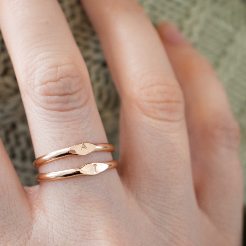 K&ouml;nnte beinhalten: Zwei goldfarbene Ringe mit einem einfachen, leicht geschwungenen Design. Der obere Ring hat den Buchstaben "A" eingraviert, und der untere Ring hat den Buchstaben "I" eingraviert.
