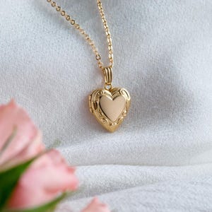 Op de afbeelding: Een goudkleurige hartvormige medaillonketting. Het medaillon heeft een sierlijk ontwerp rond de randen en een gladde hartvorm in het midden. De ketting is ook goudkleurig en delicaat. De achtergrond is een zachte, witte stof.