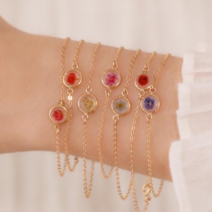 May include: Delicate gold chain bracelets, each with a small, clear, circular pendant containing a pressed flower. The flowers are in various colours, including red, pink, yellow, and blue. The bracelets are elegant.