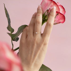 May include: A hand wearing a delicate gold chain ring with a single pearl accent, holding a pink rose with green leaves against a light pink background.
