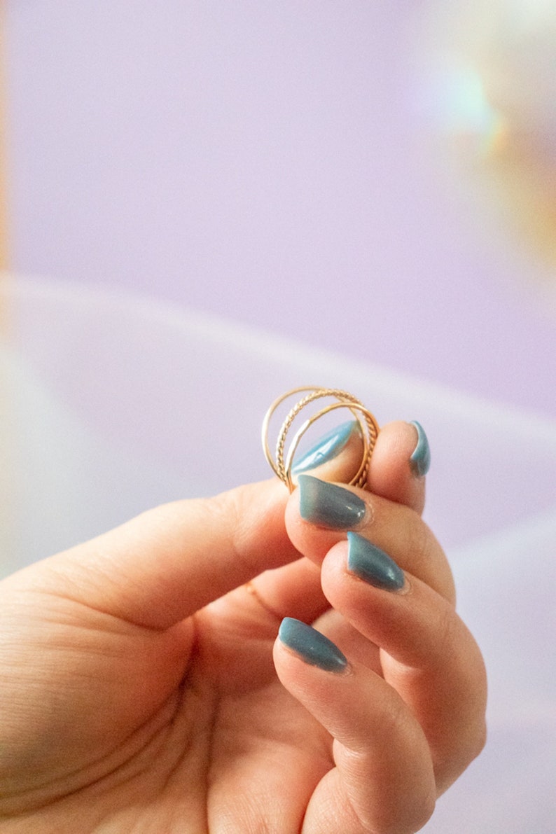 Puede incluir: Una mano con esmalte de u&ntilde;as azul sostiene un juego de tres anillos de oro. Los anillos est&aacute;n apilados uno encima del otro y est&aacute;n hechos de alambre delgado y retorcido.