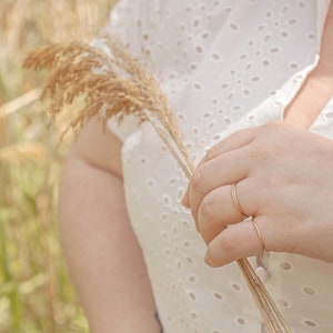Puede incluir: Una persona lleva un vestido blanco de ojal con un patr&oacute;n delicado y sostiene un tallo de hierba seca en su mano. Lleva dos anillos finos de oro en su dedo.