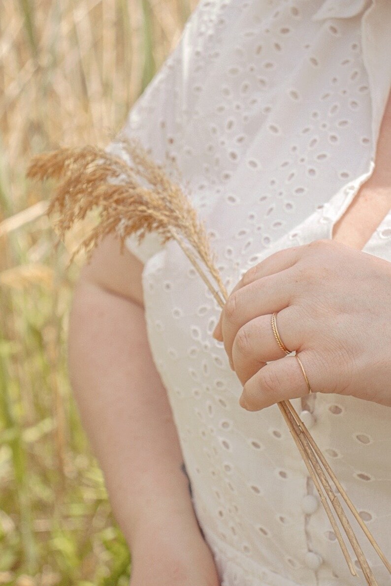 Puede incluir: Una persona lleva un vestido blanco de ojal con un patr&oacute;n delicado y sostiene un tallo de hierba seca en su mano. Lleva dos anillos finos de oro en su dedo.
