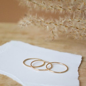 Puede incluir: Tres anillos de oro sobre un trozo de papel blanco. Los anillos son de diferentes anchos y estilos. Los anillos est&aacute;n sobre una superficie de madera.