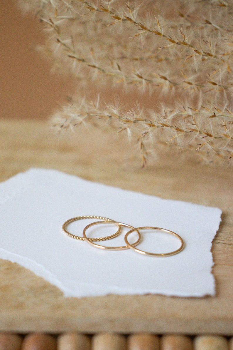 Puede incluir: Tres anillos de oro sobre un trozo de papel blanco. Los anillos son de diferentes anchos y estilos. Los anillos est&aacute;n sobre una superficie de madera.
