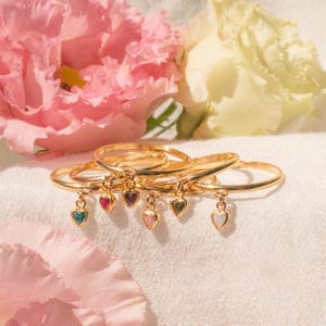 Könnte beinhalten: Goldene Ringe mit herzförmigen Anhängern in verschiedenen Farben, darunter Türkis, Rosa und Lila. Die Ringe werden auf einer weißen Oberfläche mit rosa und gelben Blumen im Hintergrund präsentiert. Die Ringe sind zart und elegant.
