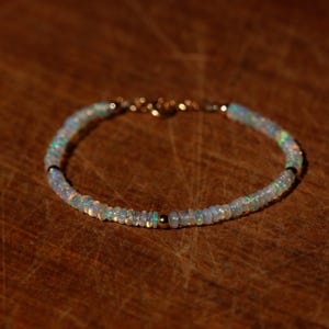 Op de afbeelding: Een delicate armband bestaande uit kleine, ronde, iriserende opaal kralen met een subtiel kleurenspel. De streng is geaccentueerd met een enkele, gladde, gepolijste goudkleurige metalen kraal en heeft een goudkleurige metalen sluiting aan de achterkant.