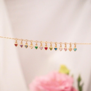 May include: A delicate gold necklace featuring a series of heart-shaped pendants, each adorned with a different colored gemstone. The gemstones include red, purple, blue, clear, green, pink, and yellow. The necklace is displayed against a soft, blurred background.