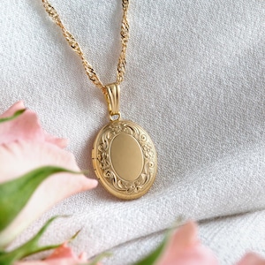Könnte beinhalten: Eine goldfarbene ovale Medaillon-Halskette mit einem verzierten floralen Design. Das Medaillon hängt an einer zarten, gedrehten Kette. Der Hintergrund ist ein weicher, neutraler Stoff, mit rosa Blumen im Vordergrund.