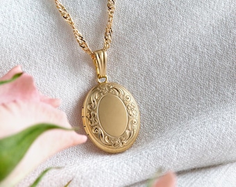 May include: A gold-colored oval locket necklace with an ornate floral design. The locket hangs from a delicate twisted chain. The background is a soft, neutral fabric, with pink flowers in the foreground.