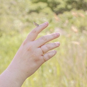 Puede incluir: Una mano con dos anillos de oro en los dedos, una peque&ntilde;a mariposa est&aacute; posada en el dedo &iacute;ndice.