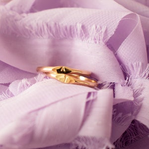 K&ouml;nnte beinhalten: Zwei goldene Ringe mit den eingravierten Buchstaben "A" und "T", die auf einem weichen, lavendelfarbenen Stoff liegen.