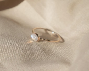 May include: A thin gold ring featuring a small, oval-shaped iridescent moonstone cabochon. The metal band is slender and polished, holding the smooth, translucent stone in a simple bezel setting that catches the light with a soft glow.