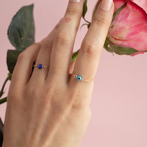 May include: Two gold chain rings, each with a small, round, blue bead. One ring has a dark blue bead, the other has a light blue bead. The rings are on a hand with a small heart tattoo and a pink rose.