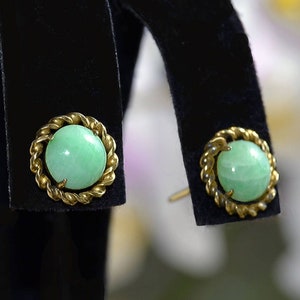 Op de afbeelding: Een paar jade oorbellen met gouden instellingen. De oorbellen zijn rond en hebben een groene jade steen in het midden. De oorbellen zijn op een zwarte fluwelen display.