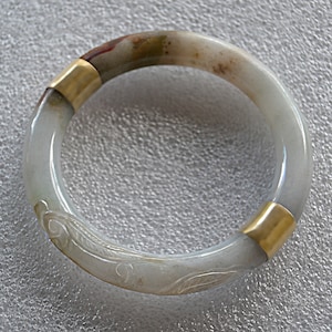 May include: A white jade bangle with gold accents and a carved floral design. The bangle is made of white jade with a light brown and green marbling effect.