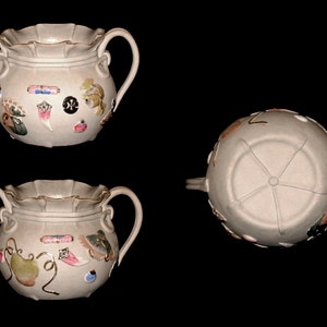 Pode incluir: Um pote de cerâmica branca com duas alças e uma borda ondulada. O pote é decorado com detalhes pintados coloridos, incluindo um peixe, uma pena, uma fita e uma pêra.