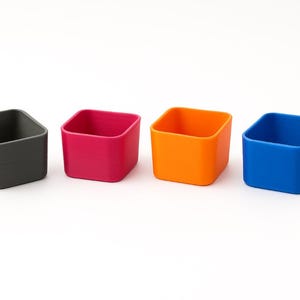 Könnte beinhalten: Vier kleine, quadratische Behälter in einer Reihe. Die Behälter sind in Grau-, Magenta-, Orange- und Blautönen gehalten. Die Behälter haben abgerundete Ecken und sind leer. Der Hintergrund ist weiß.