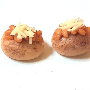 Könnte beinhalten: Zwei Miniatur-Ofenkartoffeln, belegt mit gebackenen Bohnen und geriebenem Käse. Die Kartoffeln sind hellbraun mit strukturierter Oberfläche. Die Bohnen sind rotbraun, der Käse ist weiß.