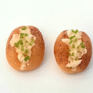 May include: Two miniature baked potato replicas, each with a golden-brown exterior and a creamy white topping, garnished with green chives. The potatoes are presented on a white surface, showcasing a detailed food miniature.