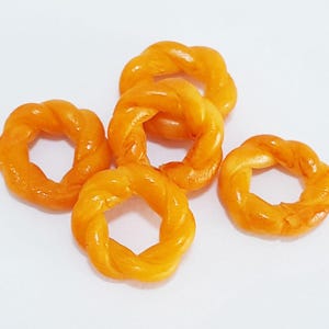 Könnte beinhalten: Fünf goldbraune, ringförmige Gebäckstücke mit einem gedrehten Design. Das Gebäck ist auf einer weißen Oberfläche angeordnet und zeigt sein glänzendes, strukturiertes Aussehen. Die Backwaren haben eine warme, einladende Farbe.