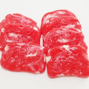 May include: A close-up of six pieces of raw meat, arranged in a pile. The meat is a vibrant red color with white marbling throughout. The pieces are rectangular and appear to be cut from a larger piece of meat.