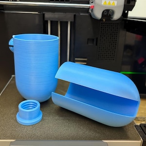 May include: A light blue 3D-printed mug with a handle, a separate lid, and a curved container. The objects are on a dark surface, with a 3D printer in the background. The mug and container have a textured surface.