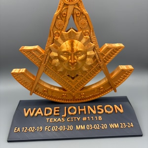 May include: A gold-colored Masonic symbol with a sun face in the center, resting on a black base. The base has the name "WADE JOHNSON" and other text. The symbol is intricately detailed.