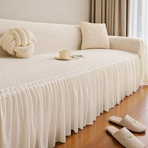May include: A cream-colored sofa with a pleated skirt, adorned with a knot-shaped pillow and a square cushion. A cup and saucer sit on the sofa, and a pair of beige slippers with embroidered details rest on the wooden floor.