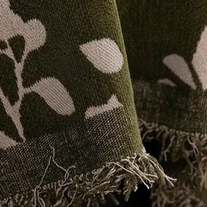 May include: Close-up of a green woven throw blanket with a white floral pattern. The blanket has a fringed edge and a textured appearance, suitable for home decor.