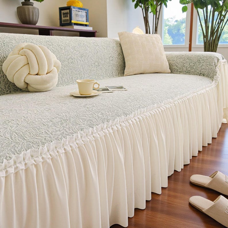 May include: A light green sofa with a white lace pattern and a white pleated skirt. A cream-colored knot pillow and a square pillow are on the sofa. A teacup and saucer sit on the sofa, along with a small magazine.