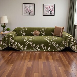 May include: A green sofa is covered with a floral patterned throw. The throw features white branches and flowers. A brown pillow with a decorative border and a round decorative pillow are on the sofa. The sofa sits on a hardwood floor.