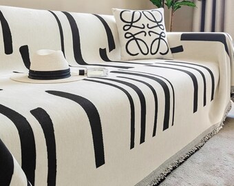 May include: A cream-colored sofa throw blanket featuring bold black vertical and curved line patterns, draped over a sofa. A white throw pillow with a black four-leaf logo design and a straw hat with a black ribbon band rest on the blanket.
