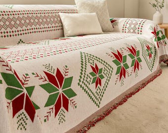 May include: A cream-colored sofa throw blanket featuring a repeating pattern of red and green geometric snowflakes and diamond shapes, finished with a multi-colored fringed edge. Two solid cream-colored throw pillows rest on the sofa.