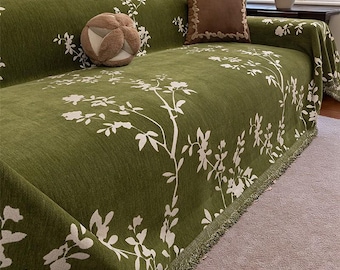 May include: A green sofa cover with a white floral pattern. The cover is draped over a sofa and features a fringe trim along the edges. A decorative pillow with a brown border and a round, textured pillow are on the sofa.