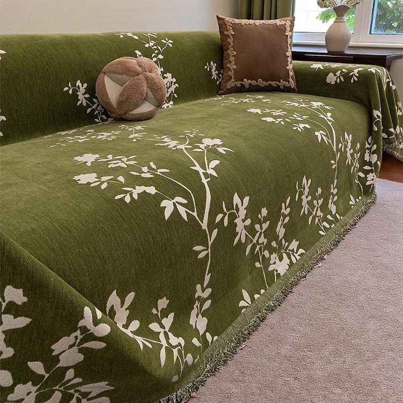 May include: A green sofa cover with a white floral pattern. The cover is draped over a sofa and features a fringe trim along the edges. A decorative pillow with a brown border and a round, textured pillow are on the sofa.