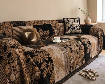 May include: A sofa covered in a black and tan patterned throw blanket featuring floral and geometric motifs. A matching square pillow, a round brown textured pillow, and a white teacup sit on the seat. A pair of plaid slippers rests nearby.