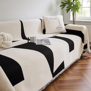 May include: A cream-coloured sofa is draped with a black and cream geometric patterned throw blanket. A matching pillow with a minimalist face design rests on the sofa. A decorative knot pillow, a striped mug, and a magazine are also on the sofa.