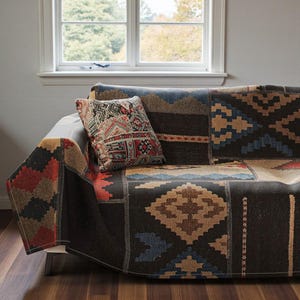 以下が含まれることがあります： 黒、茶色、赤、青の色合いの幾何学模様が特徴のパッチワークデザインのソファ。同様の模様の装飾枕がソファに置かれています。ソファは窓の近くに配置されています。