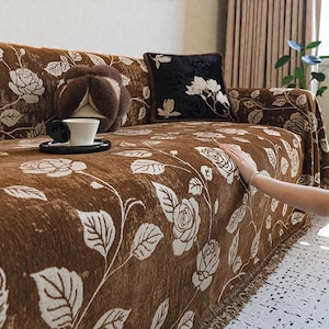 May include: Brown sofa cover with a white floral pattern. The cover features a fringe along the bottom edge and is draped over a sofa. A black pillow with white floral embroidery and a brown decorative pillow are also visible.