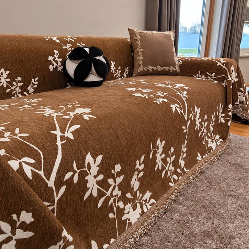 May include: Brown sofa cover with a white floral pattern. The cover has a fringed edge and is draped over a sofa. A decorative black and white pillow and a brown square pillow with a decorative border are on the sofa.